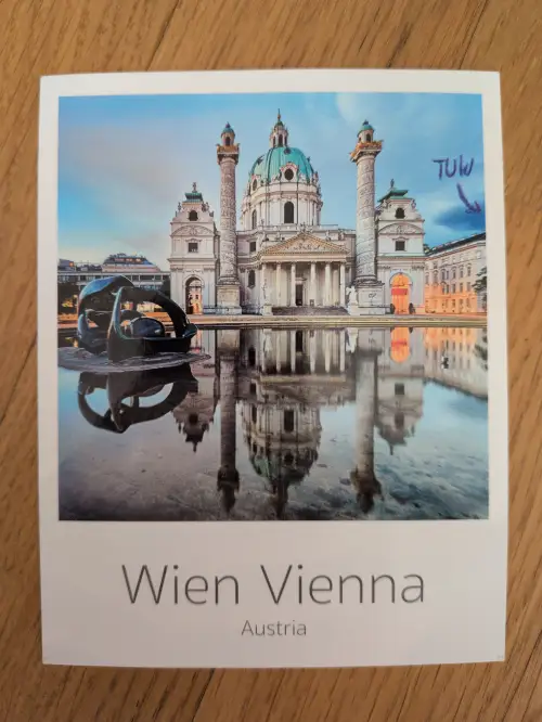 Ein Postkartenbild der Wiener Karlskirche mit einer Skulptur im Vordergrund, die sich beide im Wasser spiegeln, und dem Text "Wien Vienna Austria".