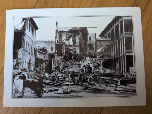 Ein Schwarz-Weiß-Foto zeigt eine zerstörte Stadtstraße voller Schutt und Trümmer, mit teils eingestürzten Gebäuden und einigen Menschen, die sich darin bewegen.