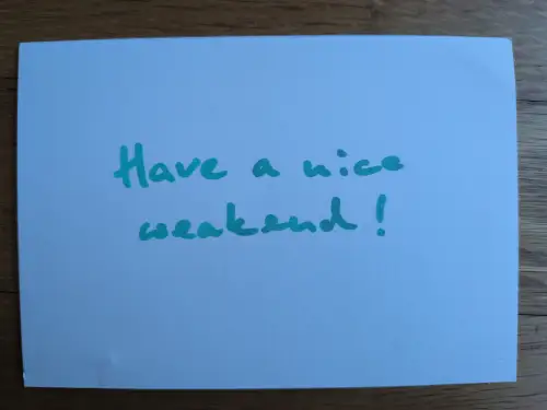 Eine hellblaue Karte mit der türkisfarbenen Aufschrift "Have a nice weekend!" liegt auf einem Holzhintergrund.