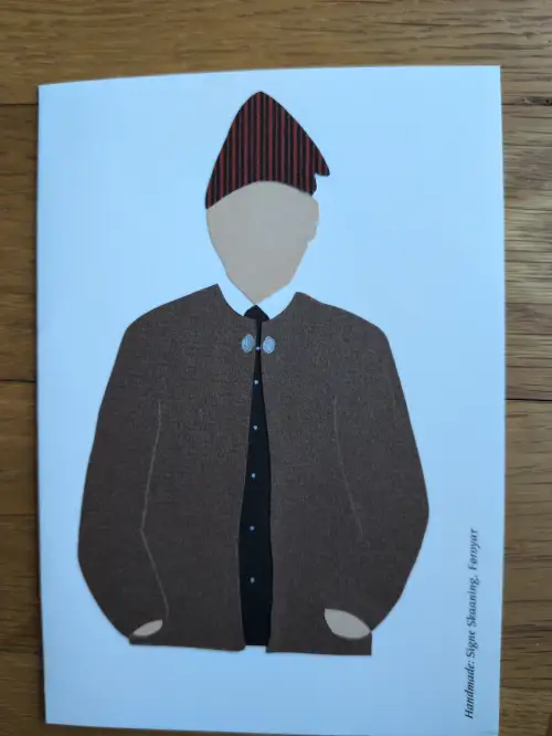 Ein Papierschnitt zeigt eine männliche Figur in brauner Tracht mit einem rot-schwarz gestreiften Hut und silbernen Spangen auf hellblauem Grund.