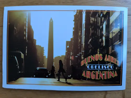 Das Bild zeigt den Obelisken von Buenos Aires in Argentinien, eingerahmt von dunklen Hochhäusern, mit schemenhaften Menschen und dem Schriftzug "BUENOS AIRES OBELISCO ARGENTINA".