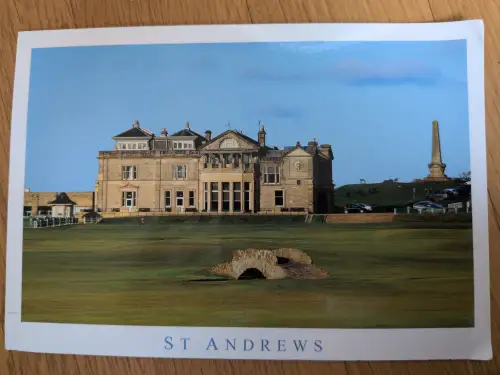Das Bild zeigt den Golfplatz von St Andrews mit einem großen historischen Gebäude, einer kleinen Steinbrücke im Vordergrund und einem Obelisken auf einem Hügel unter blauem Himmel.