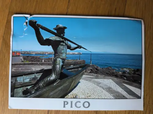 Eine grünlich-braune Statue eines Fischers mit Hut und Harpunenstab steht in einem Boot vor blauem Meer und Himmel.