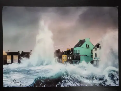 Gewaltige, hoch aufpeitschende Wellen brechen sich dramatisch direkt vor einer Reihe bunter Küstenhäuser unter einem stürmischen Himmel.