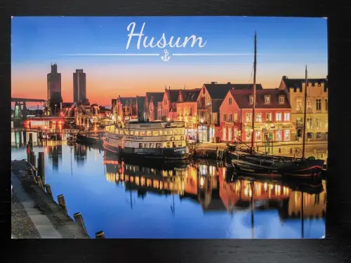 Eine Postkarte zeigt den Husumer Hafen in der Abenddämmerung mit beleuchteten Schiffen und historischen Häusern, die sich im Wasser spiegeln, unter dem weißen Schriftzug „Husum“.